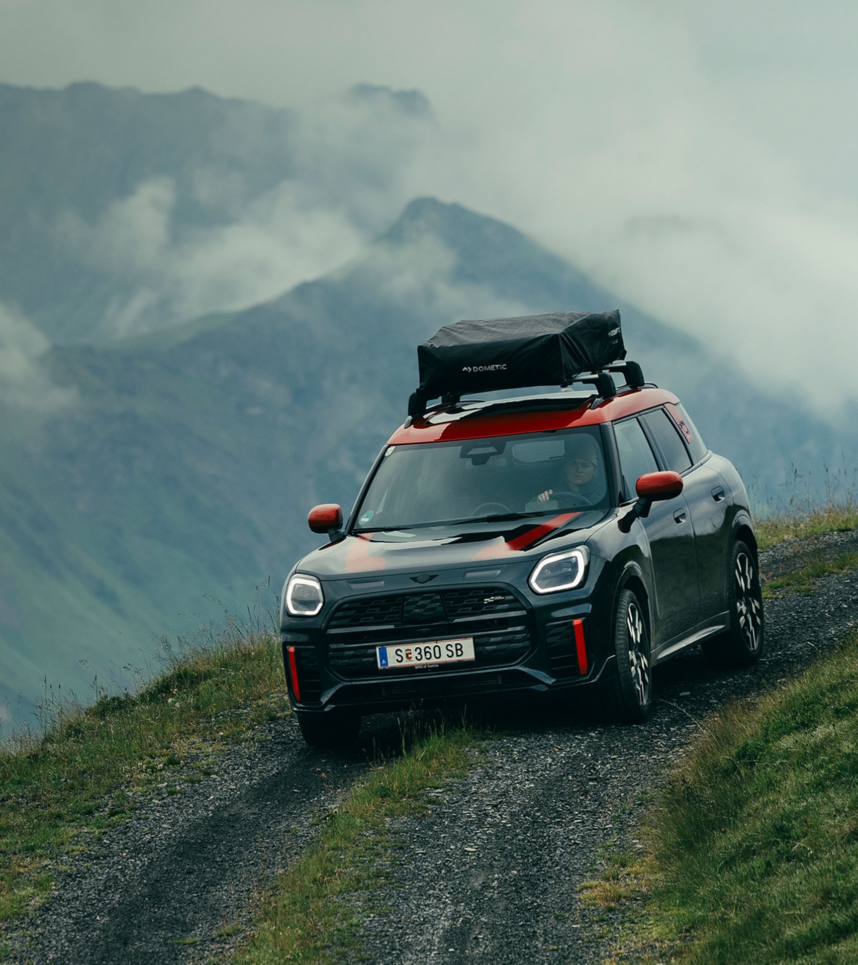 The new MINI John Cooper Works Countryman ALL4 with roof rack pictured driving around a corner on an unpaved mountain road, with a steep drop-off and misty mountain views in the background.